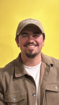 a man in a hat and jacket smiling in front of a yellow background